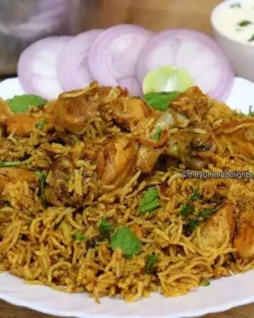 close-up of pressure cooker chicken biryani served on a white plate. Garnished with onion rings and lemon wedge. Served with Raita.