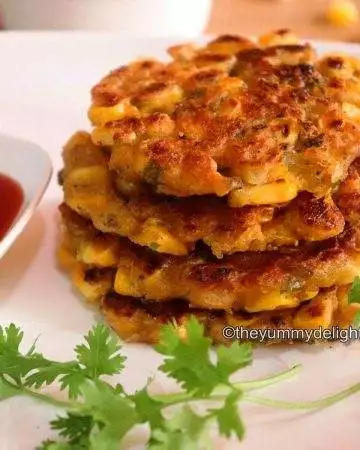 Corn fritters stacked & served in a white plate. Served with tomato sauce. Garnished with coriander leaf.