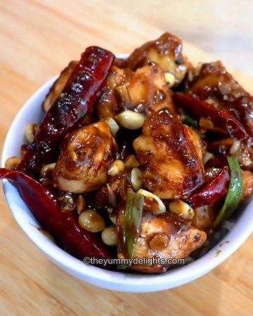 Kung pao chicken served in a white bowl. Garnished with some peanuts & spring onion greens.
