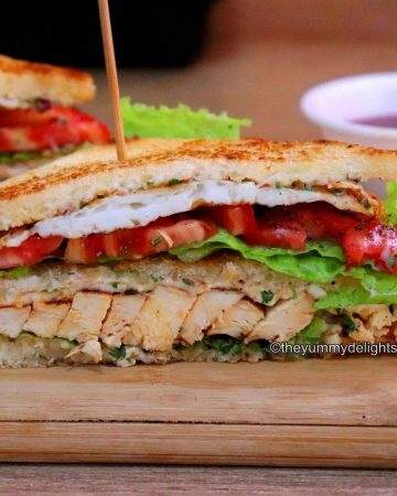 Close-up view of chicken club sandwich. The club sandwich is placed on a chopping board. THe layers of grilled chicken, tomato, cheese slice, lettuce leaves and fried eggs are visible.