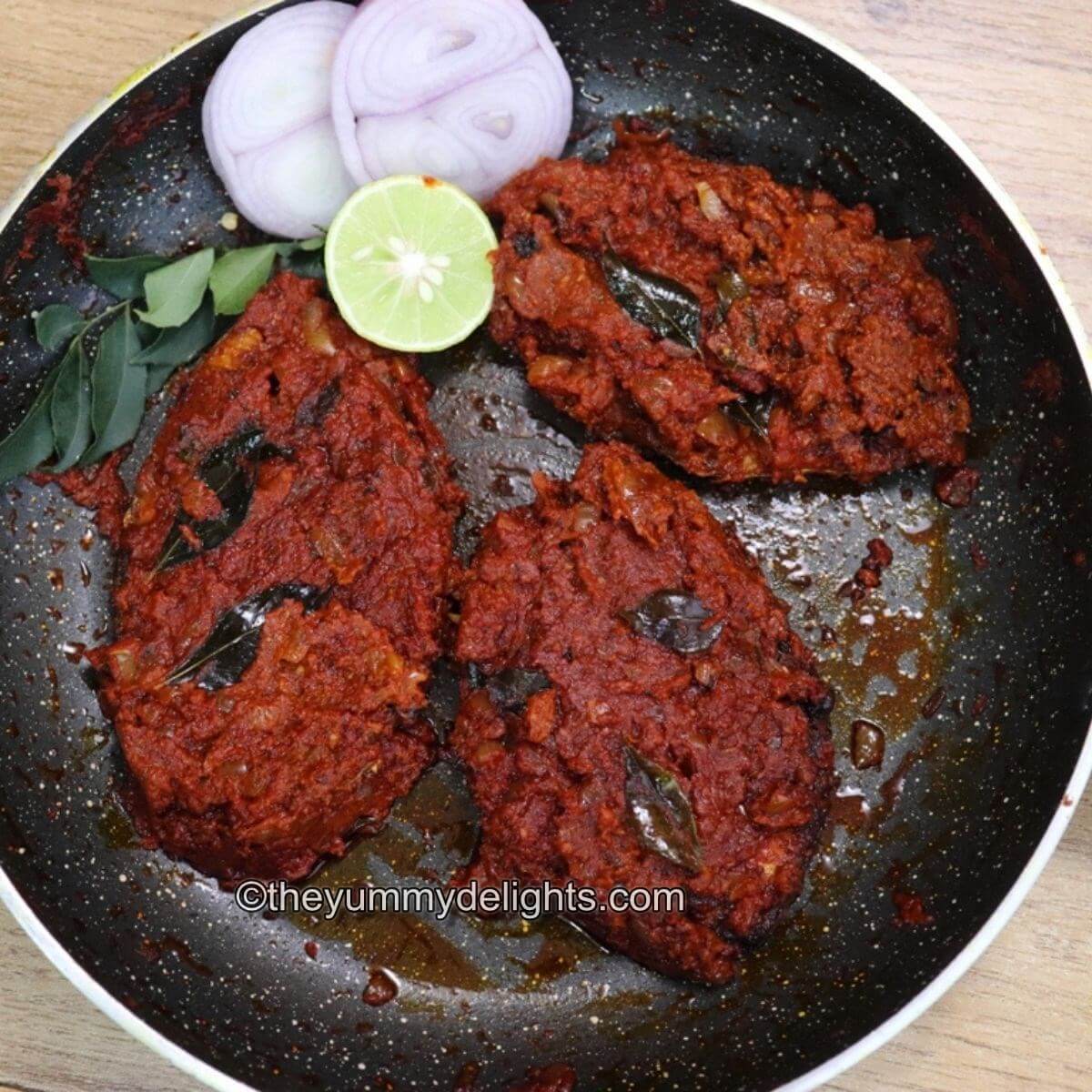 Clode-up of fish masala fry in a pan with onion slices, lemon wedge and curry leaves.
