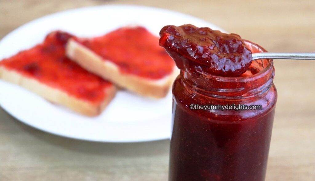 spoon full of strawberry jam placed on a strawberry jam jar. Two bread slices with jam are placed on the side on this jar.