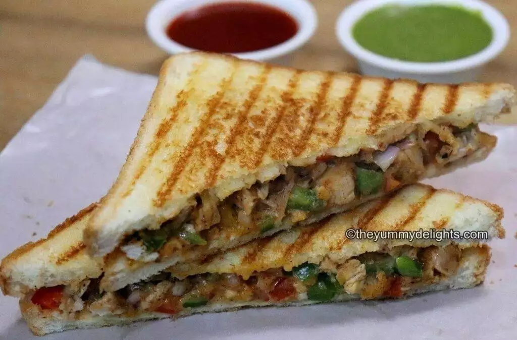 side view of grilled chicken sandwich served with tomato sauce and green chutney on the side.