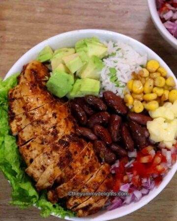 close-up of chicken fajita rice bowl. It shows fajita bowl with rice as base and topped with lettuce leaves, chicken, avocado, corn, beans, salsa and cheese.
