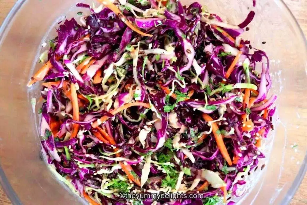 Top view of Greek yogurt coleslaw in a large glass bowl. You can see colorful cabbage, carrots and cilantro. Garnished with roasted sesame seeds and flax seeds.
