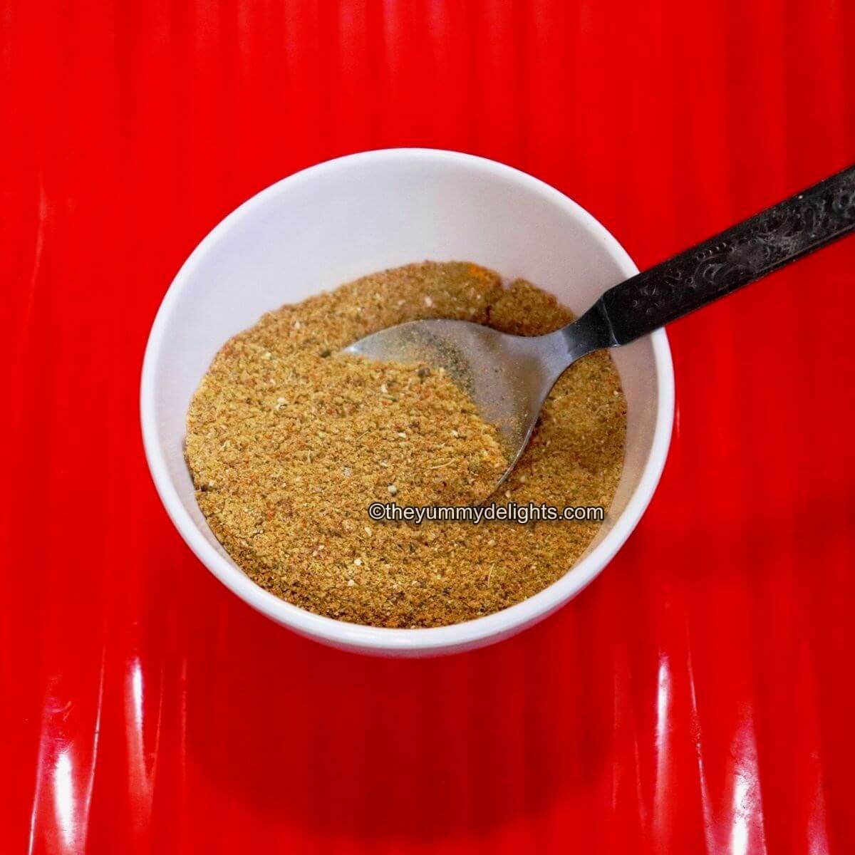 close-up of homemade curry powder in a white colored bowl with a spoon in it. The bowl is placed on a red colored plate.