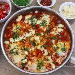 close-up of shakshuka in a skillet. It is garnished with cheese and cilantro. Served with salad and cheese on the side.