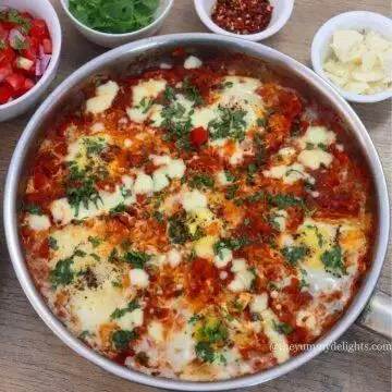 close-up of shakshuka in a skillet. It is garnished with cheese and cilantro. Served with salad and cheese on the side.