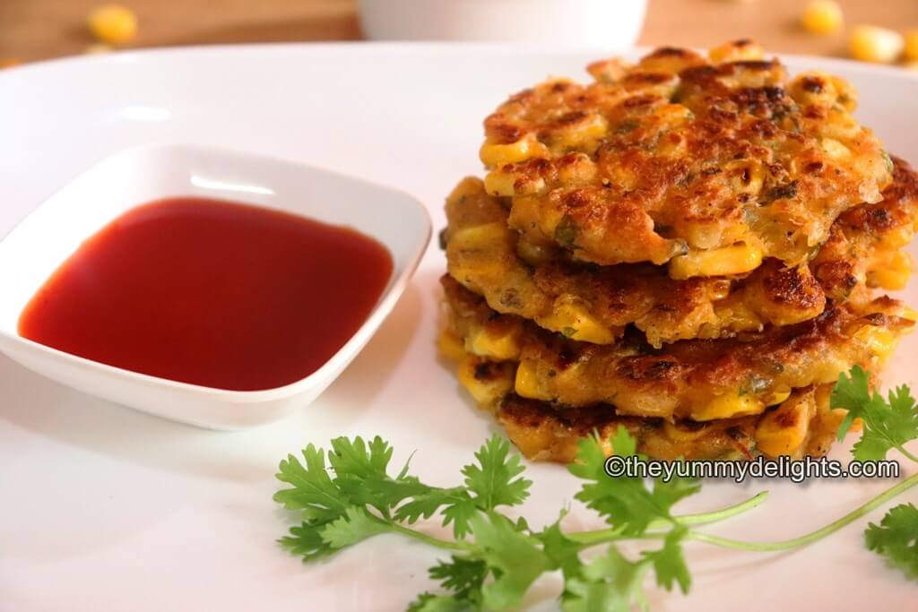 crispy corn fritters stacked on a white colored plate. Served with tomato ketchup on the side.