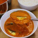 a ladle full of sardine curry is held over a bowl of sardine curry.