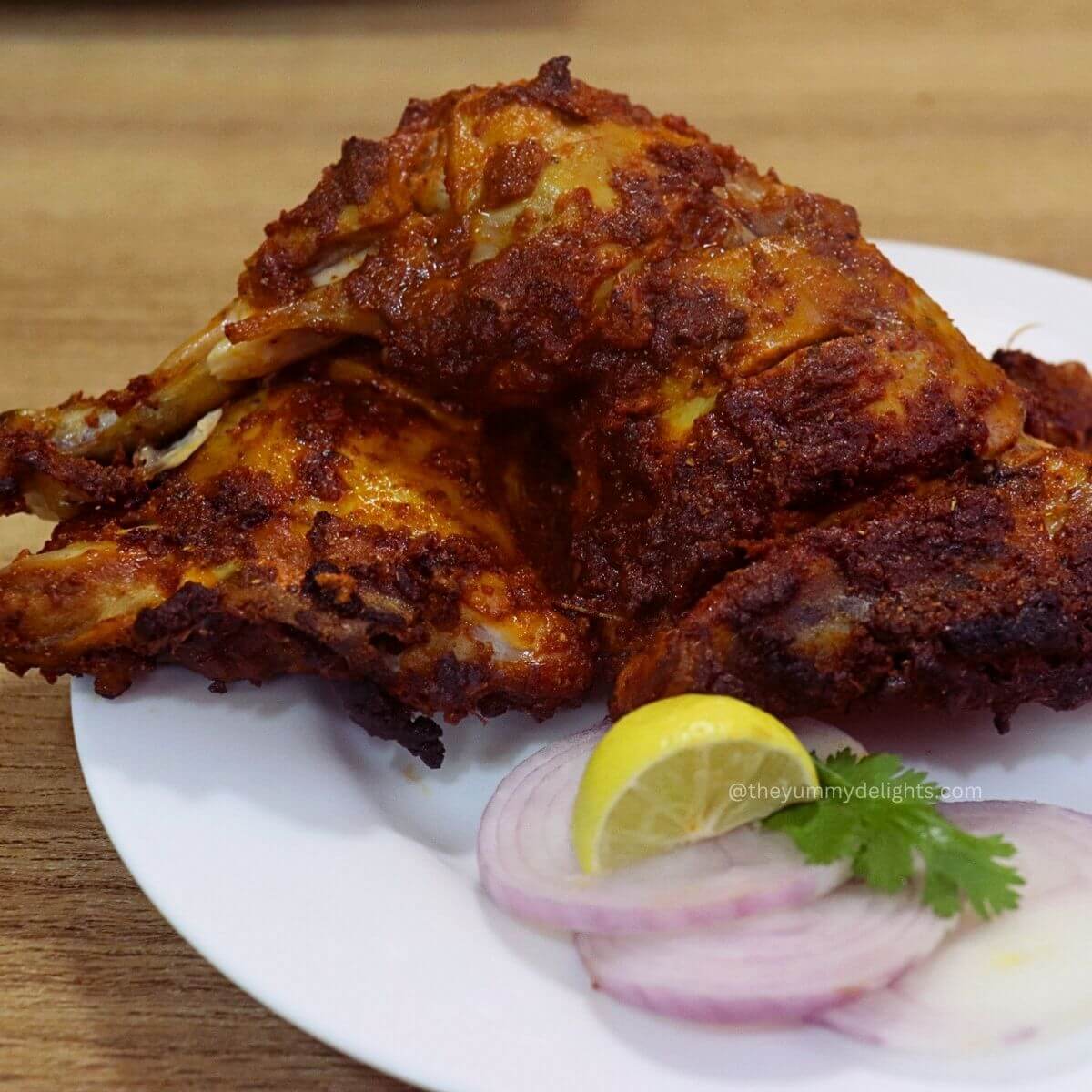 close-up of baked tandoori chicken placed on a white colored plate, served with onion slices and a lemon wedge.