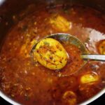 close-up of dhaba-style egg curry in a ladle, held over a rustic pot of spicy onion tomato masala and garnished with fresh coriander.