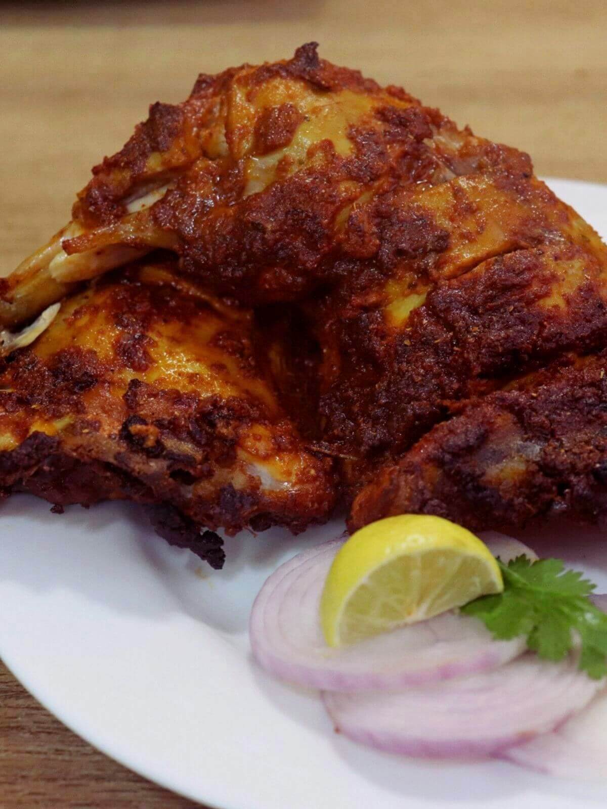 close-up of baked tandoori chicken served on a plate with onion slices and lemon wedge served on the side.