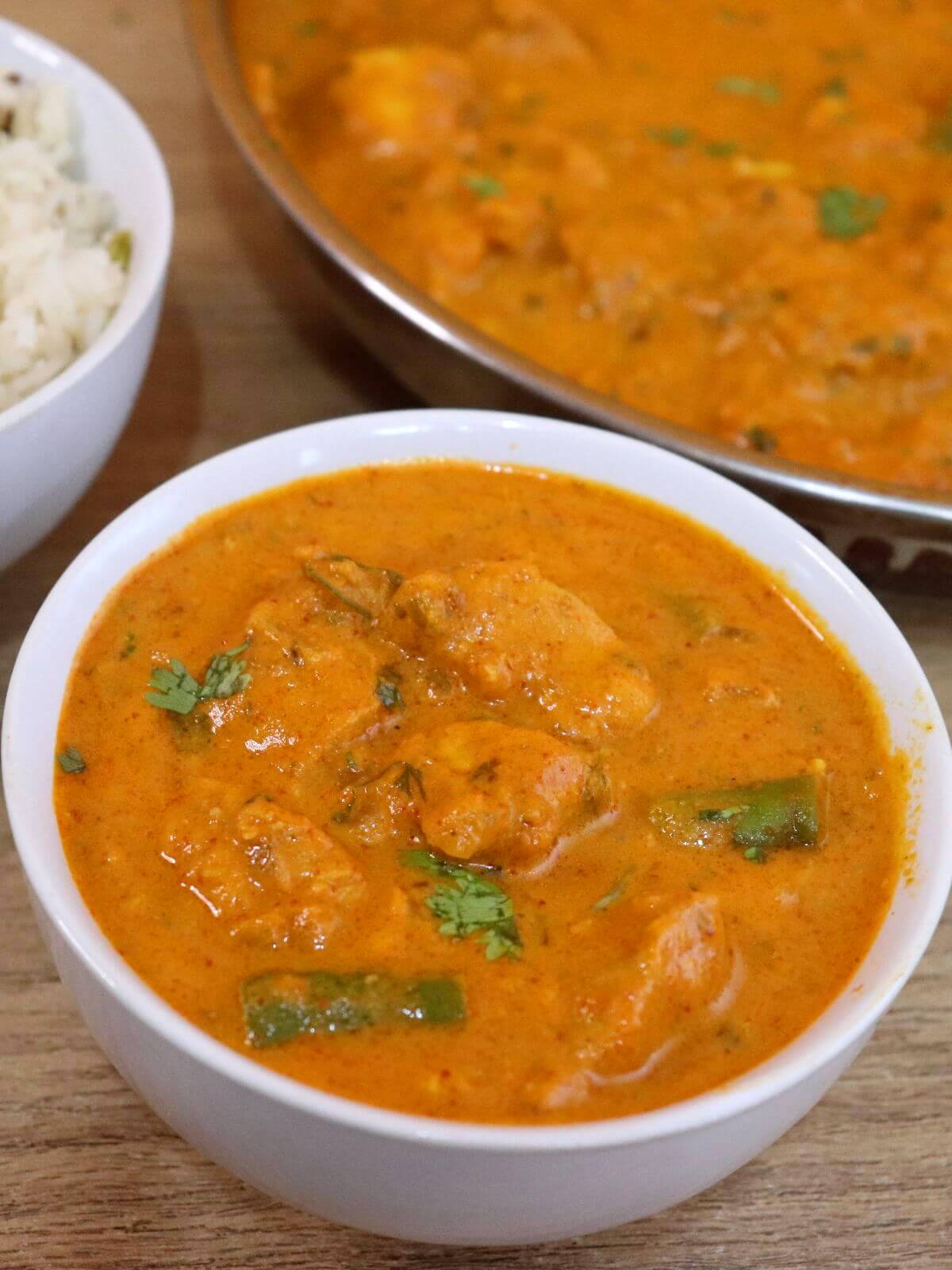 An up-close view of boneless chicken curry served in a bowl. It is served with rice served in a separate bowl on the side, and a skillet containing more chicken curry.