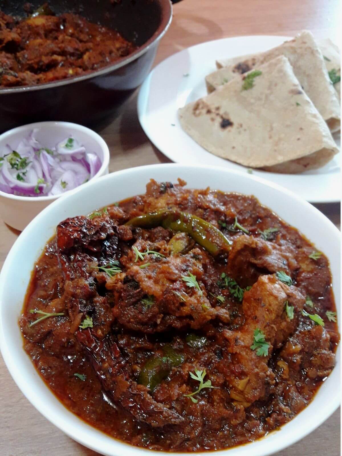 close-up of chicken bhuna masala. It is served with a bowl full of salad, roti served in another plate and more curry in a kadhai on the side.