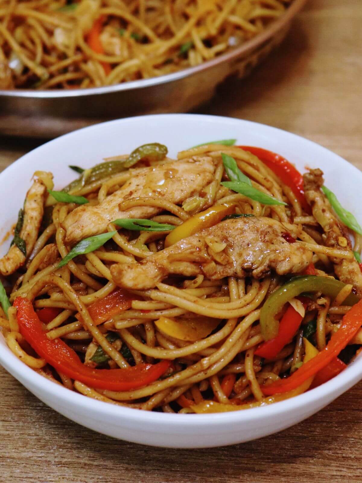 chicken stir fry with noodles are served on a white bowl. A pan full of more noodles kept on the side.