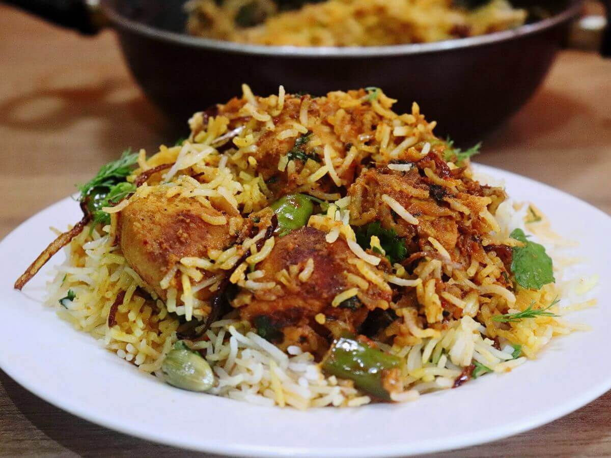 chicken tikka biryani is served on a white plate, with a pot full of more biryani kept on the side.