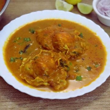 Close-up shot of Mangalorean egg drop curry with two eggs on a serving plate. Served with some onion slices and lemon wedge on the side.