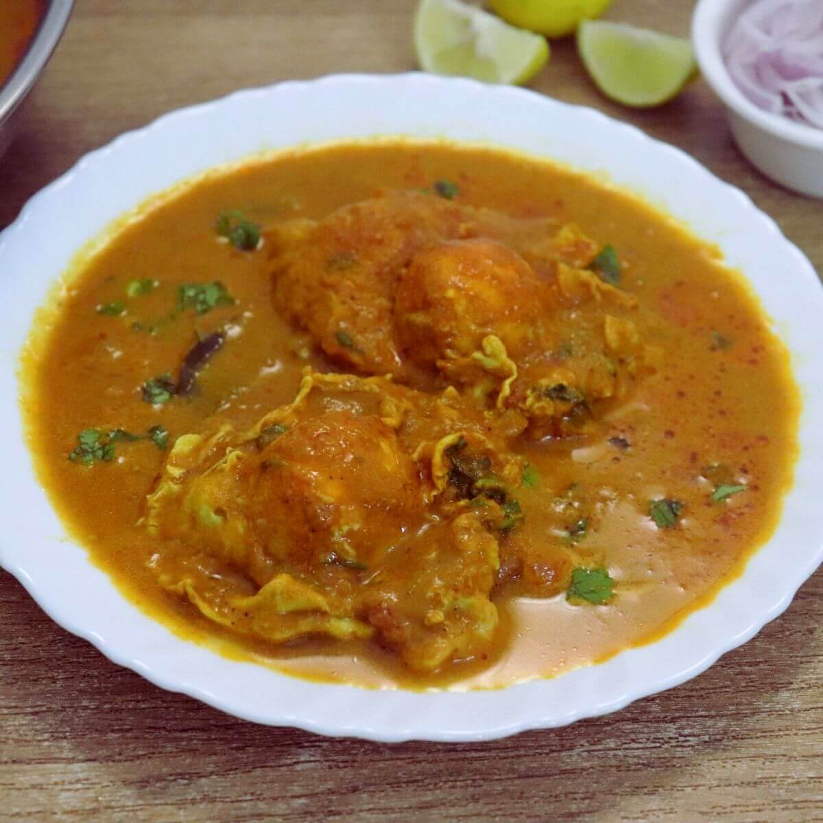 Close-up shot of Mangalorean egg drop curry with two eggs on a serving plate. Served with some onion slices and lemon wedge on the side.