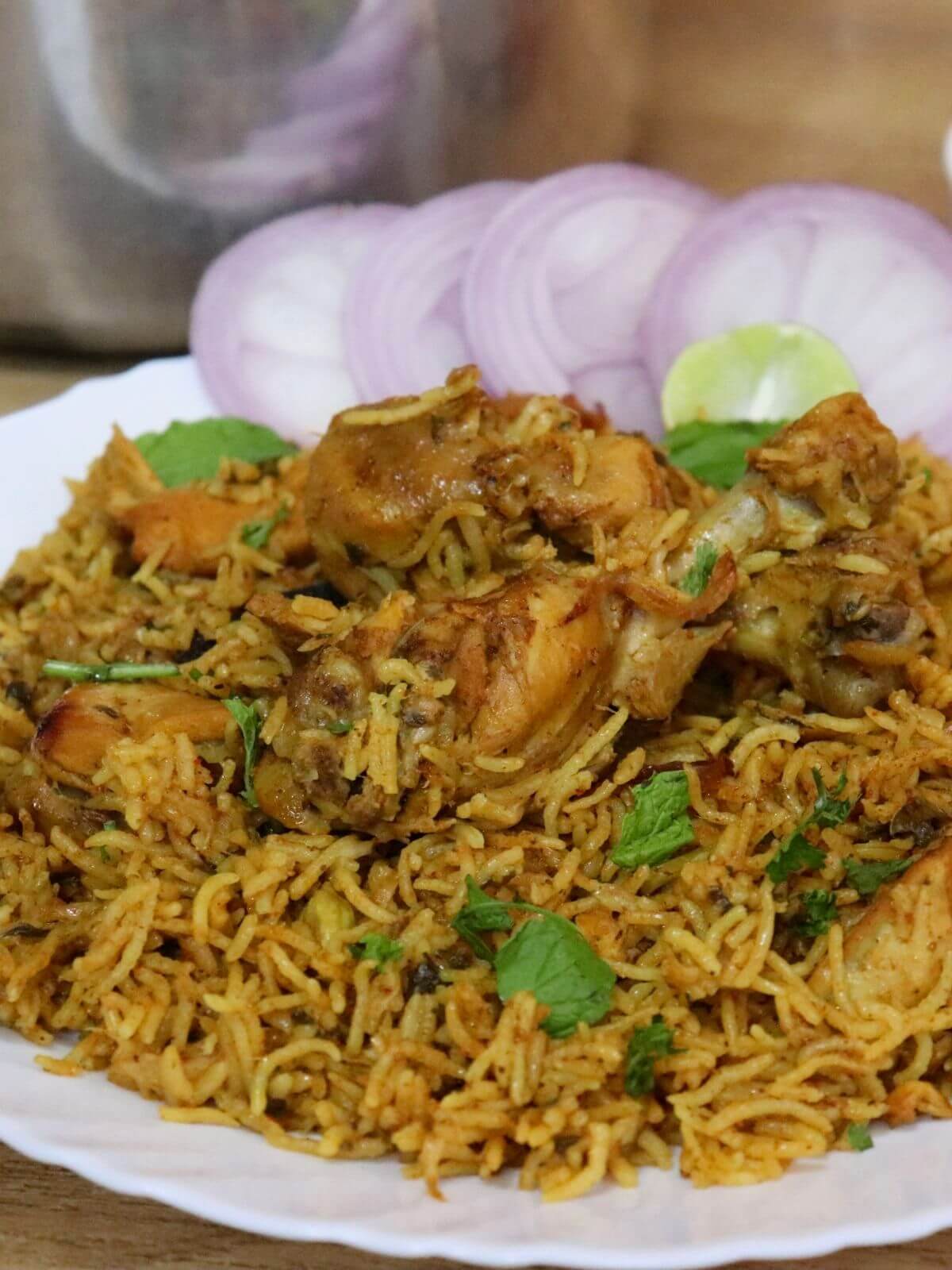 A tight close-up of pressure cooker chicken biryani, served with onion rings, and lemon wedge. It is garnished with coriander leaves.