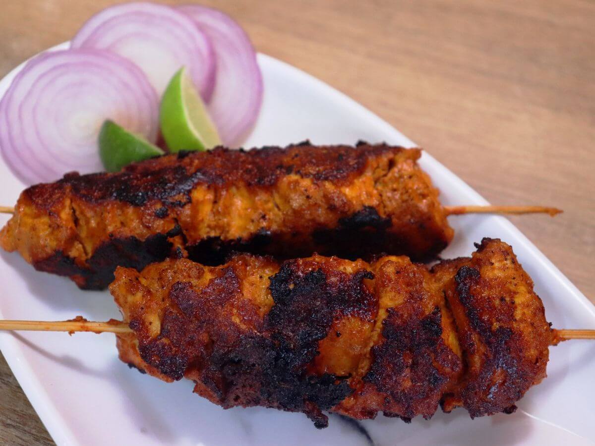 close-up of boti kebab in a skewer served on a plate with onion slices and a lemon wedge on the side.
