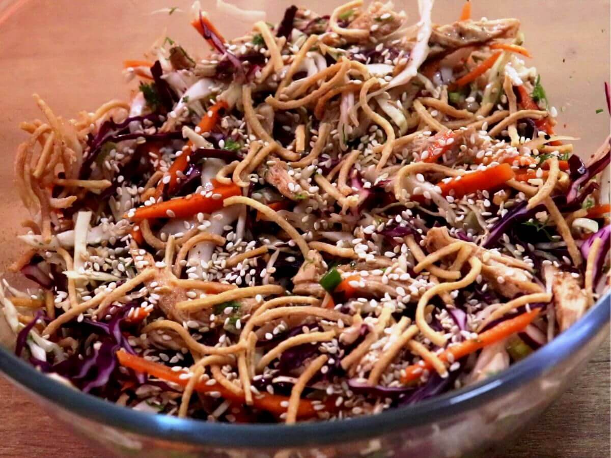A close-up shot of a tossed chicken salad, generously topped with crispy fried noodles and toasted sesame seeds.