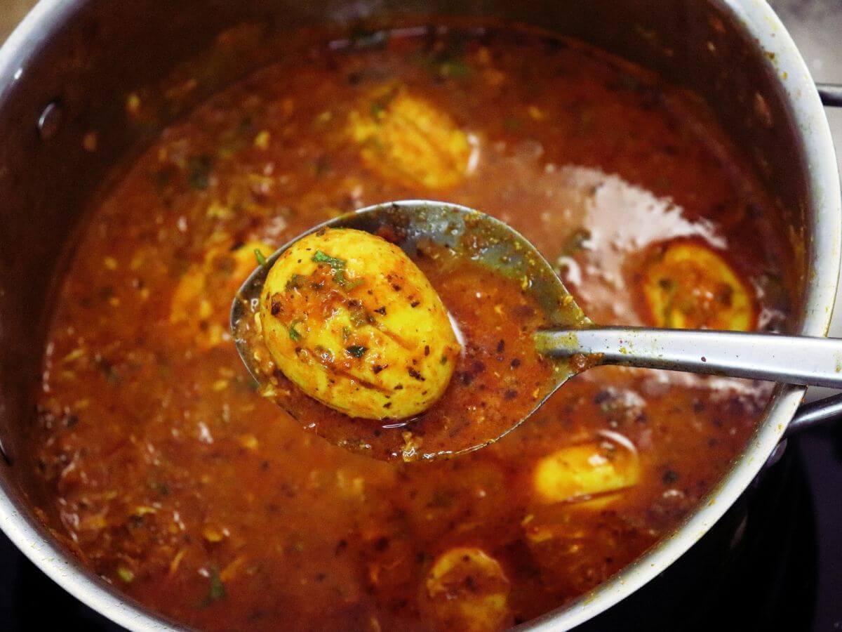A close-up of dhaba style egg curry is shown, with a ladle, positioned over a pot and full of the egg curry.