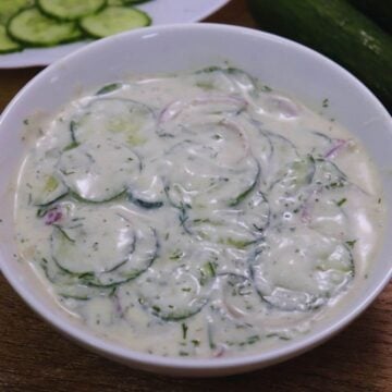 Close-up of cucumber slices coated in creamy yogurt dressing, served in a white color bowl.