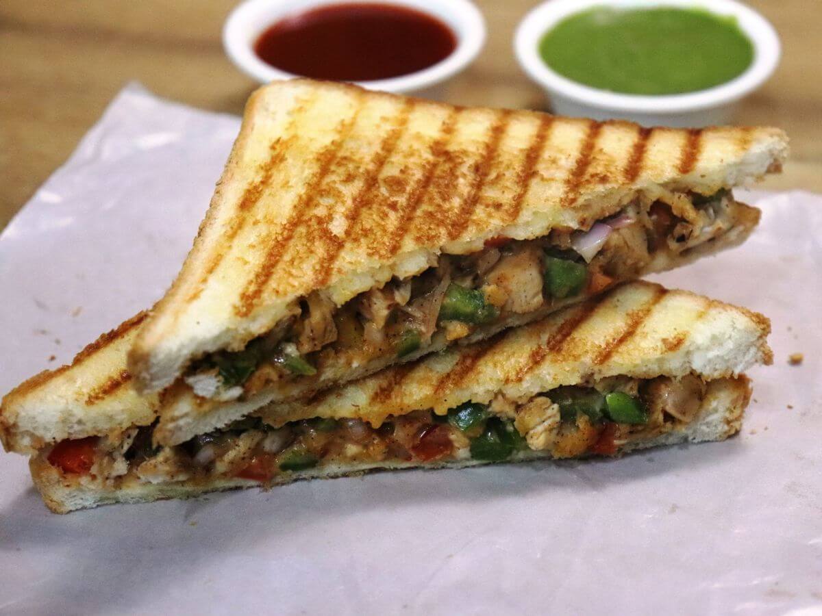 Stacked grilled chicken sandwich in a close-up, presented alongside dipping sauces of green chutney and tomato ketchup.