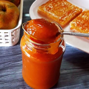 A close-up shot of homemade mango jam in a glass jar, with a spoon full of jam balanced on its rim.
