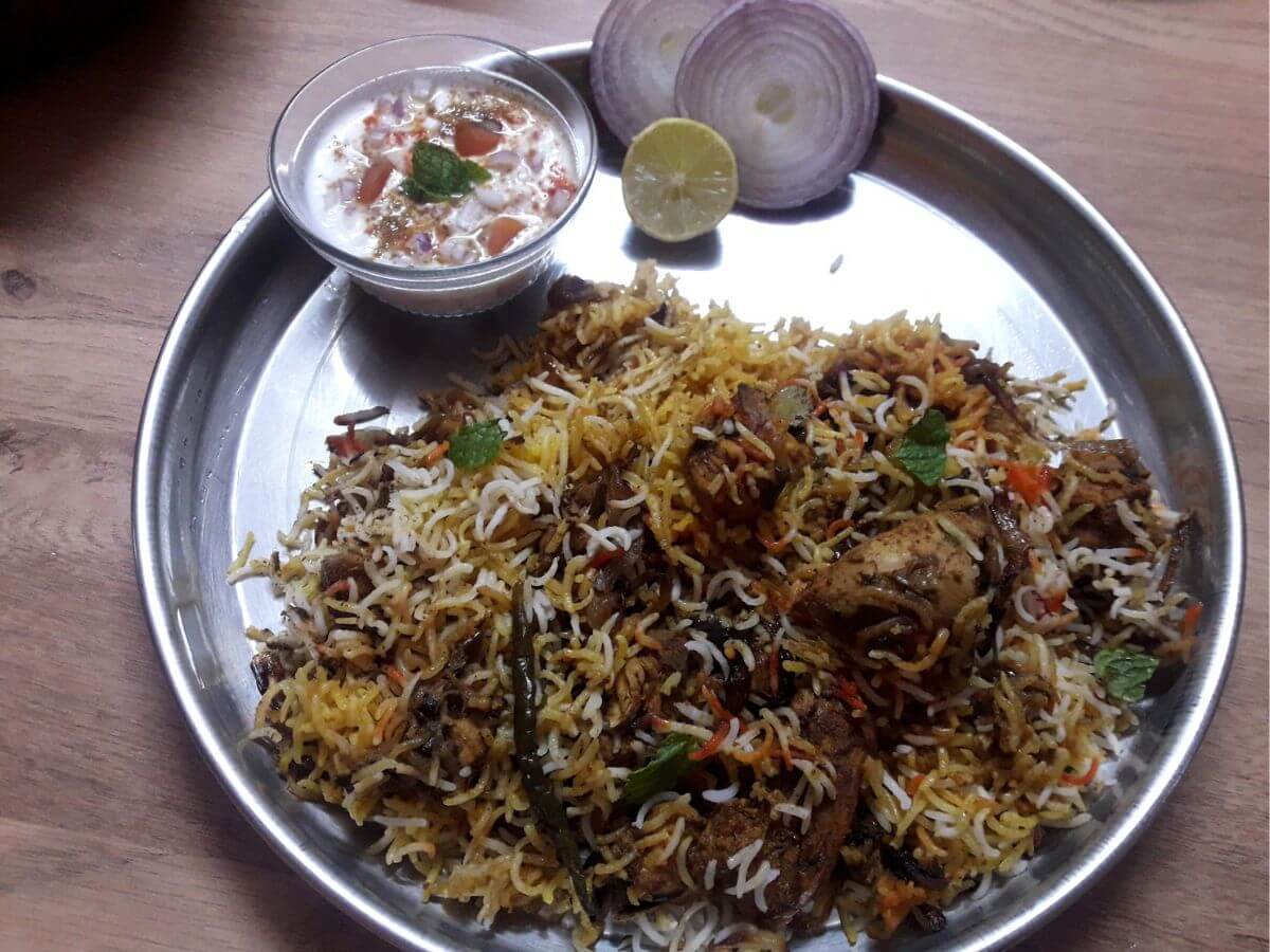 close-up of hyderabadi pakki biryani served on a plate with onion slices, lemon slice, and raita.