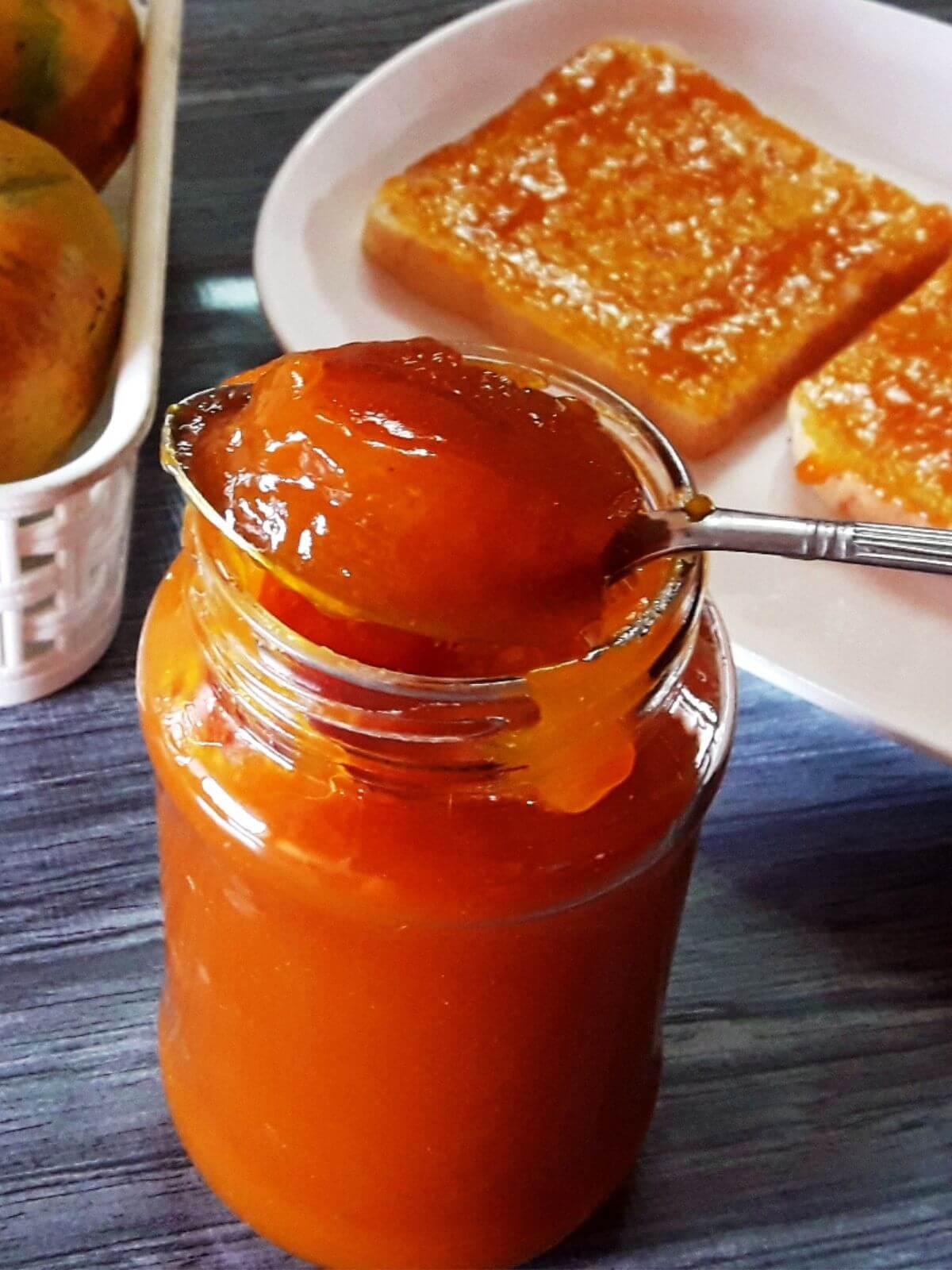 A close-up shot of homemade mango jam in a glass jar, with a spoon full of jam balanced on its rim.