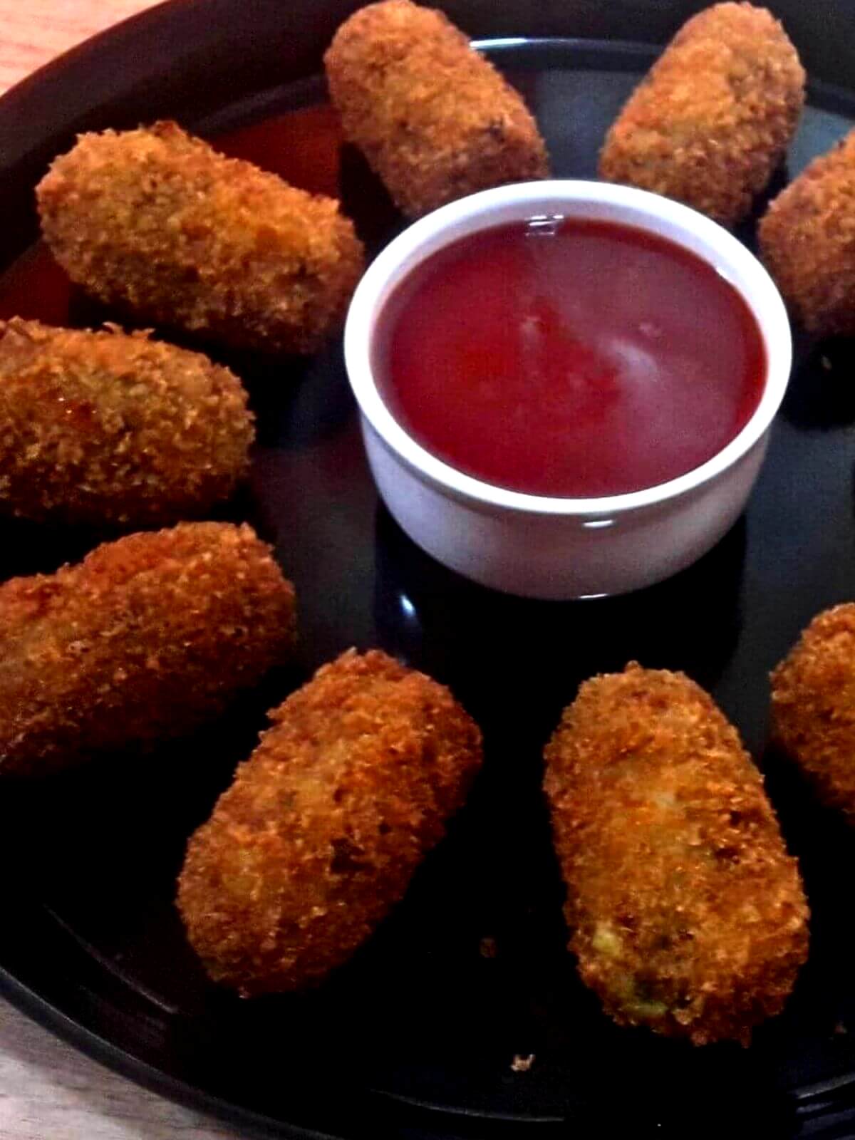 close-up of potato nuggets served in a black color plate with a small bowl of ketchup on side.