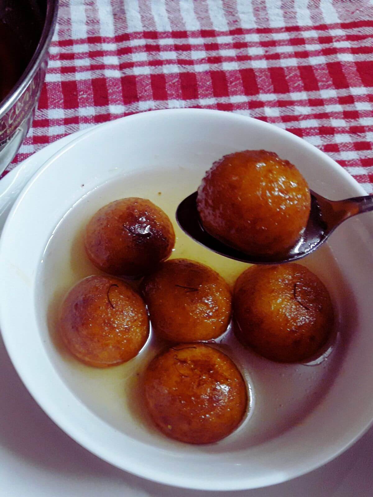 close-up of suji ke gulab jamun in a bowl. There are 5 gulab jamun in a bowl, and one is on a spoon held over the bowl.