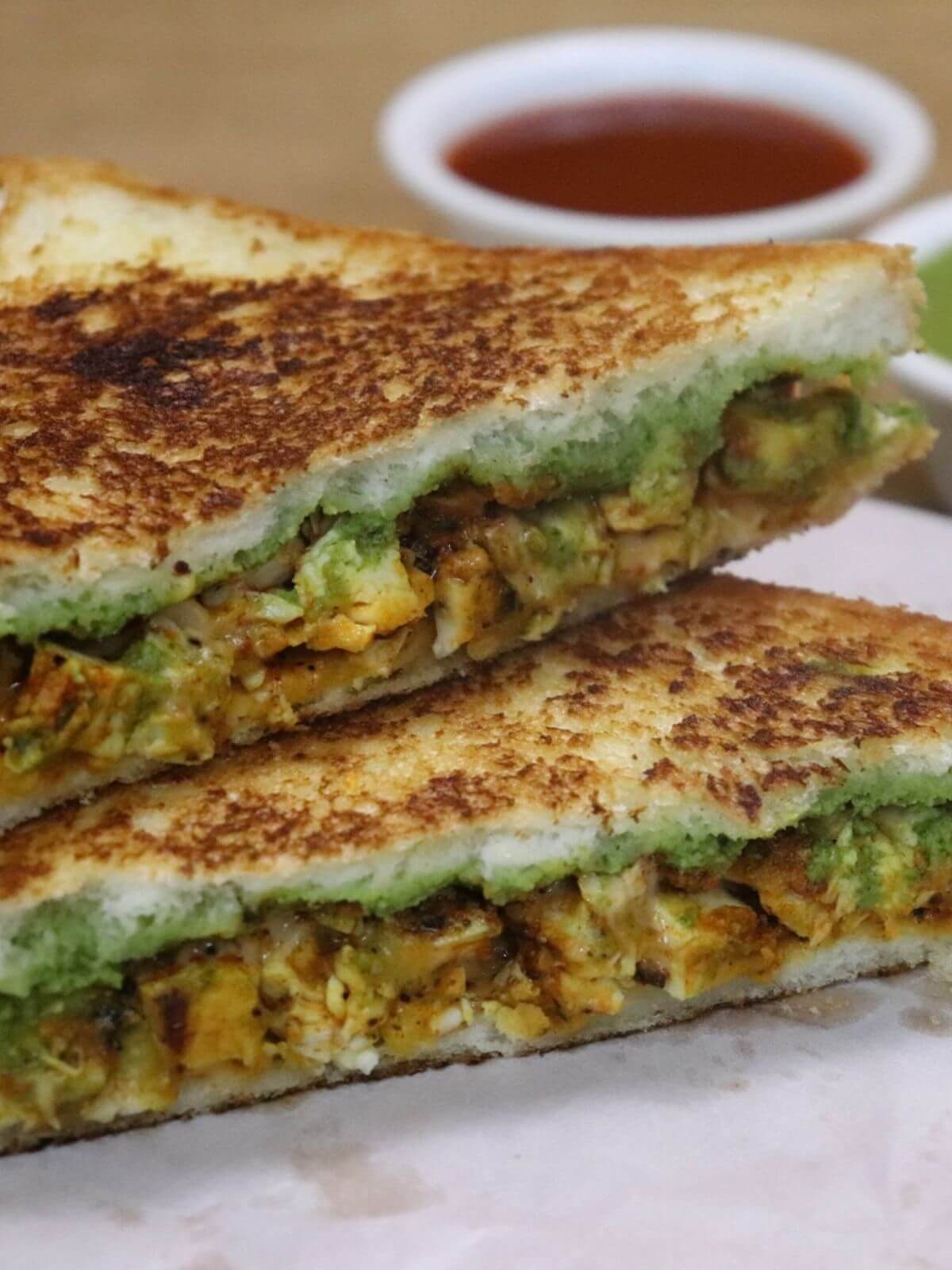 A golden-brown tandoori chicken sandwich, cut in half and showing its layers, presented on a piece of butter paper.