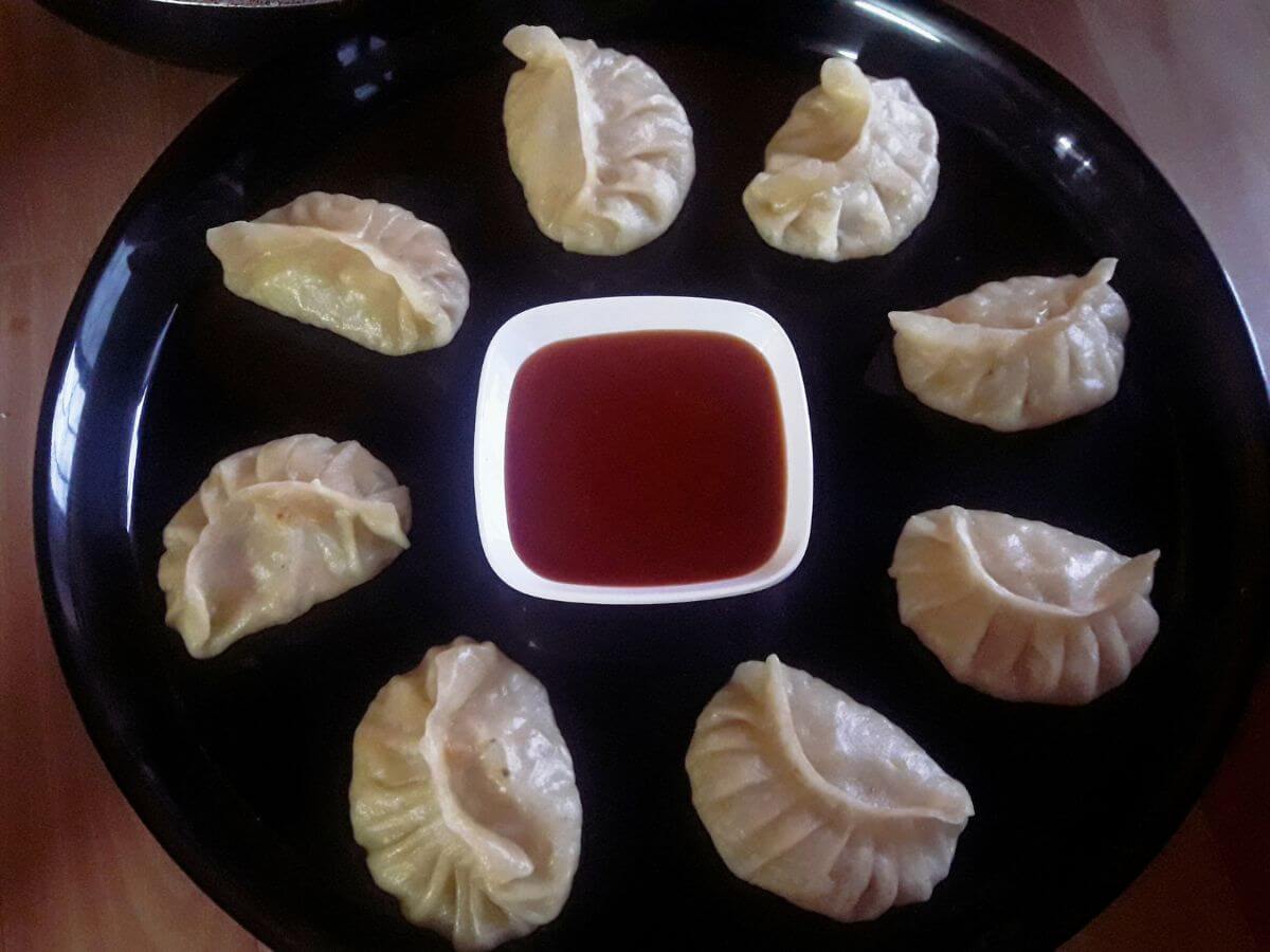 A close-up shot of vegetable momos served on a black plate with a small bowl of tomato ketchup.
