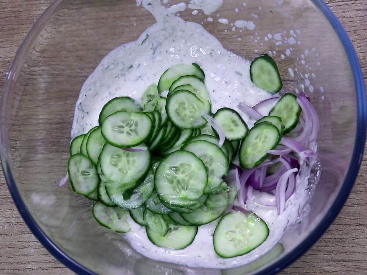Mixing cucumber and red onion with Greek yogurt dressing in a bowl.