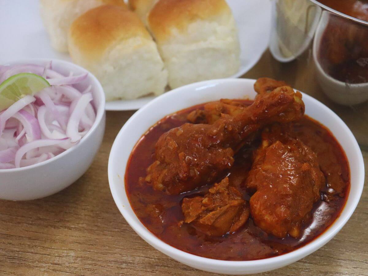 Goan chicken vindaloo served in a white bowl, accompanied by pav, onion slices, and a lemon wedge in a separate bowl.