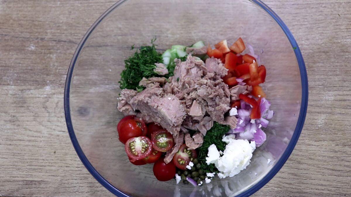 Tuna, cucumber, cherry tomatoes, red bell pepper, onion, olives, capers, fresh herbs, and feta cheese in a large bowl for Mediterranean tuna salad.