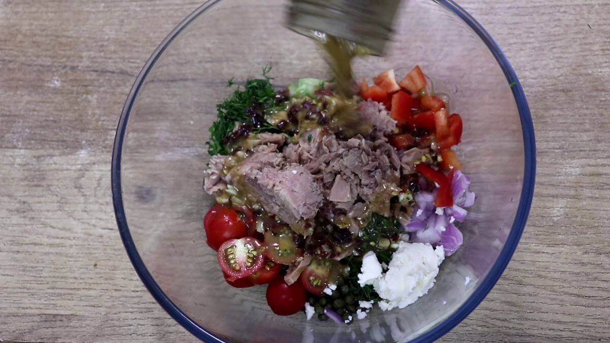 Lemon olive oil dressing being poured over Mediterranean tuna salad ingredients in a mixing bowl.