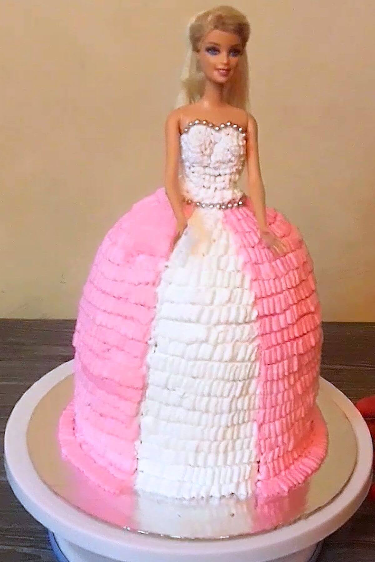 Barbie doll cake decorated with pink and white whipped cream ruffles on a silver cake board placed on a turntable.