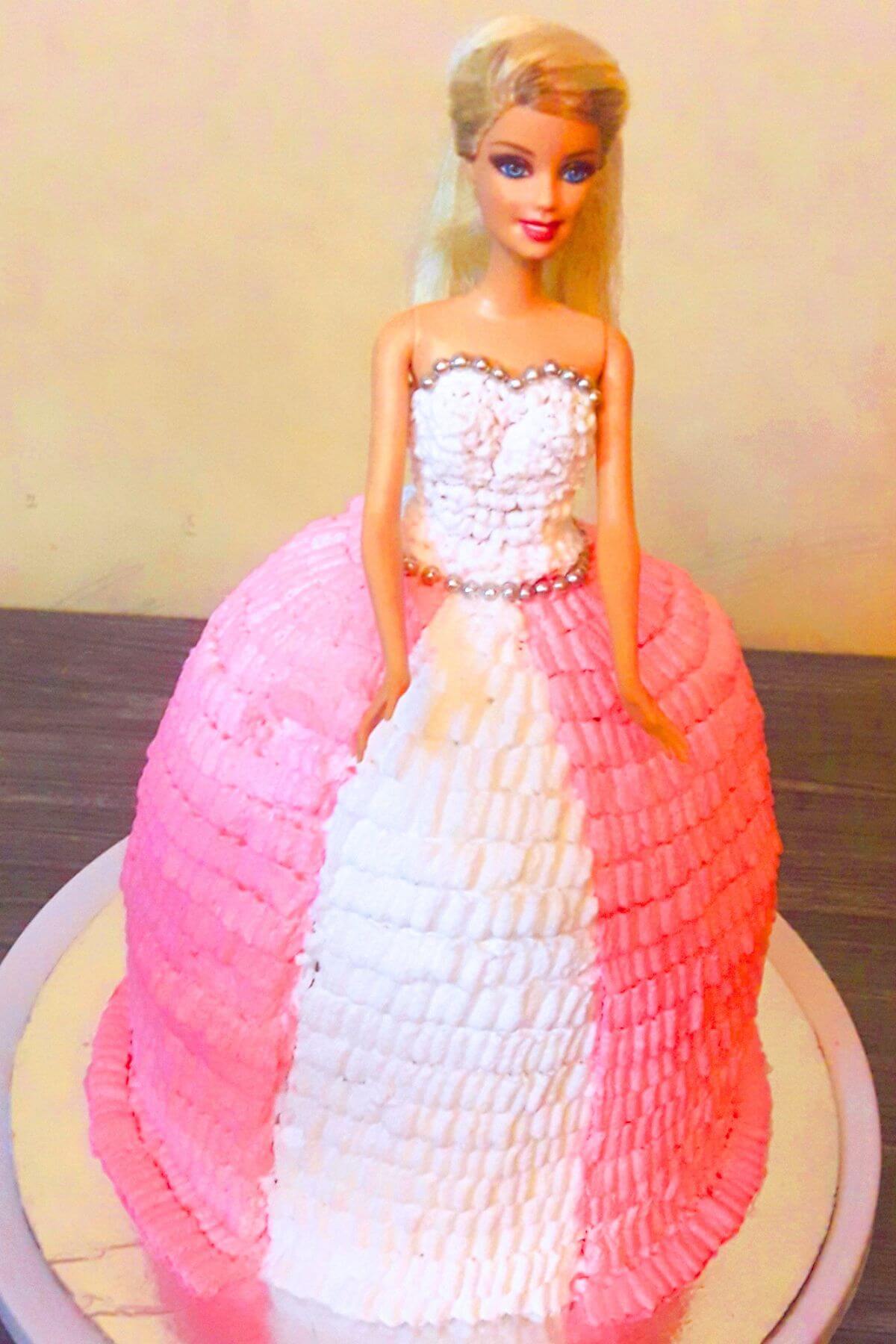 Barbie doll cake with pink and white whipped cream ruffles and edible silver pearl decorations on a silver cake board.